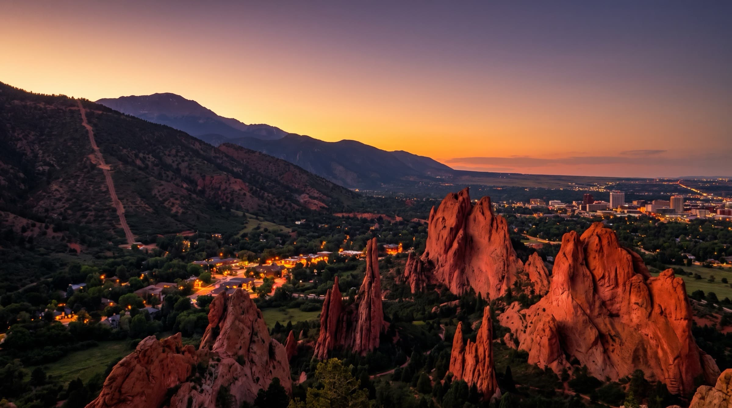 Garden of the Gods red rock formations at twilight above Manitou Springs, with Pikes Peak and downtown Colorado Springs beyond