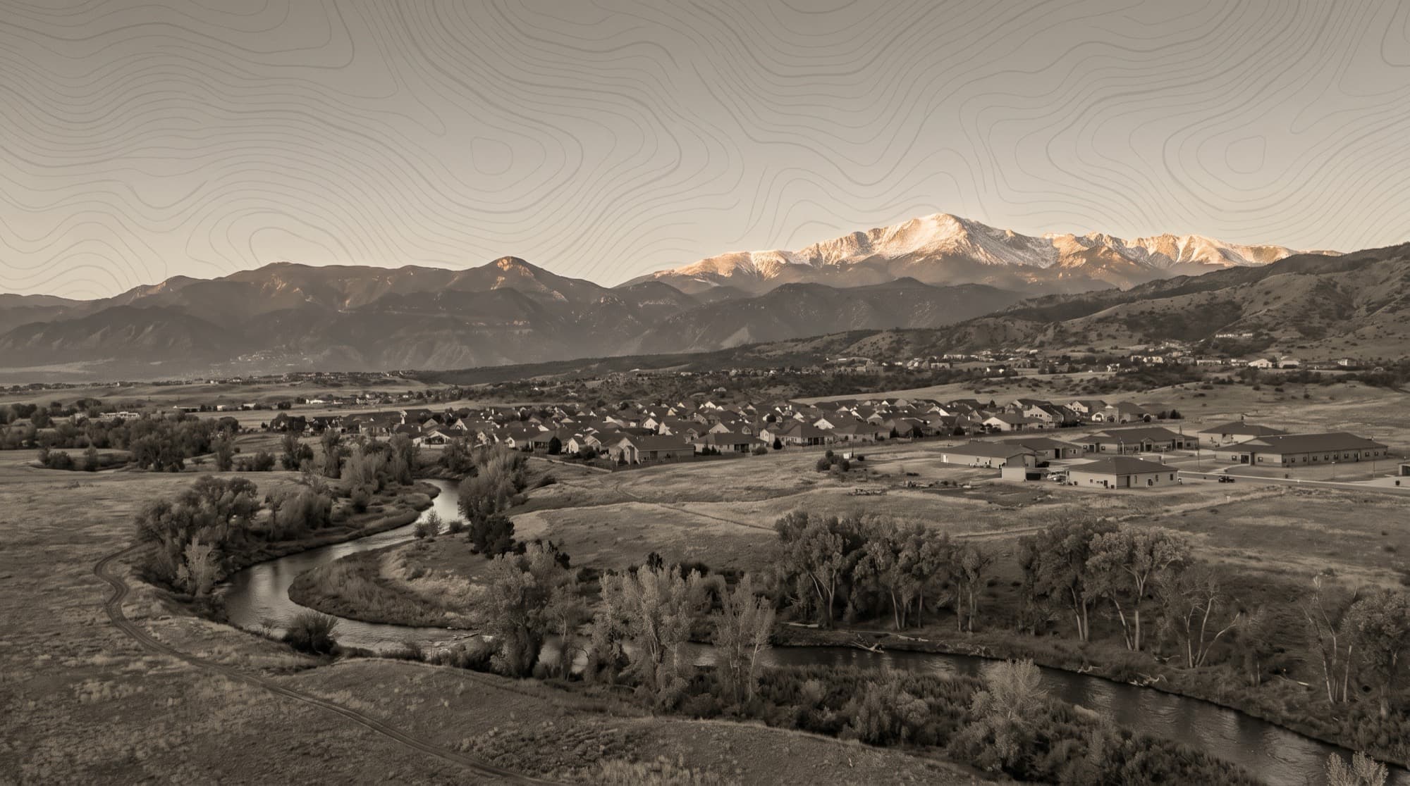 Fountain Creek winding through cottonwood bottomland below the Security-Widefield neighborhoods, with alpenglow on Pikes Peak and the Front Range to the west at dusk